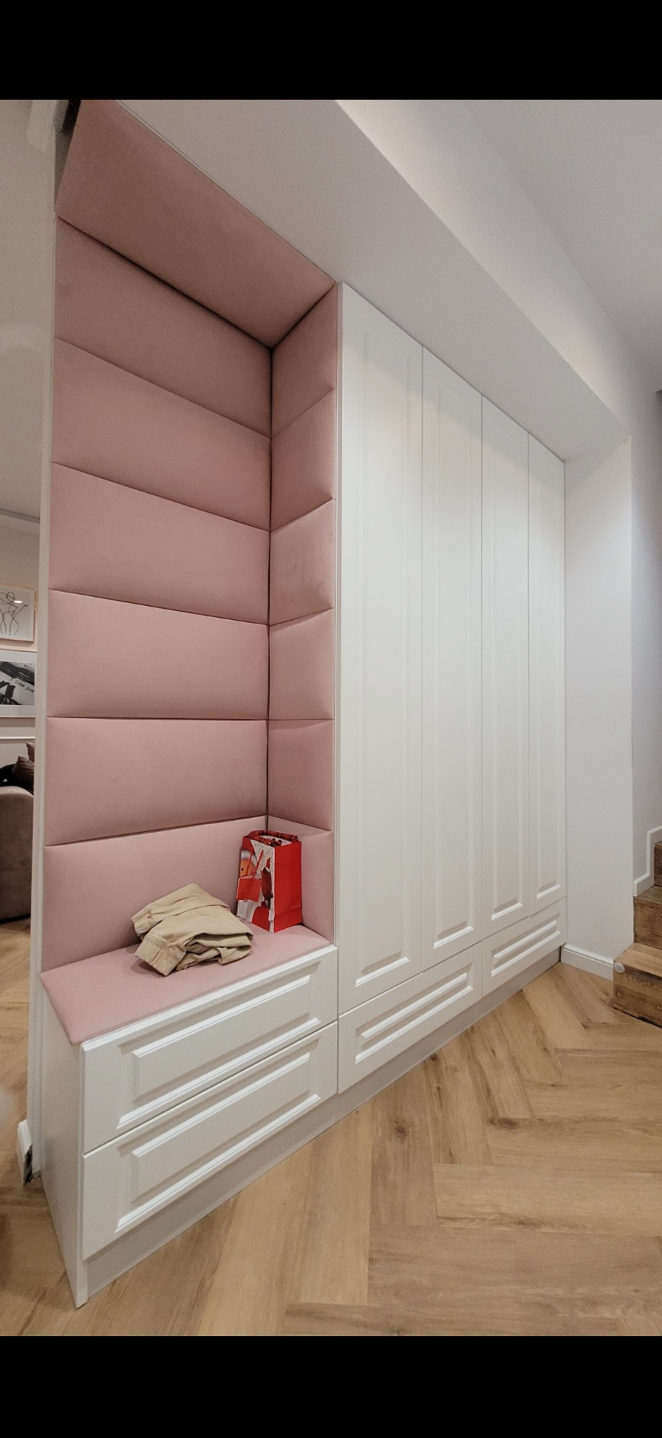 Biała szafa wnękowa z siedziskiem obitym różowym welurem. Na siedzisku leżą ubrania i czerwona torba prezentowa. Pod siedziskiem dwie szuflady. Parkiet ułożony w jodełkę.