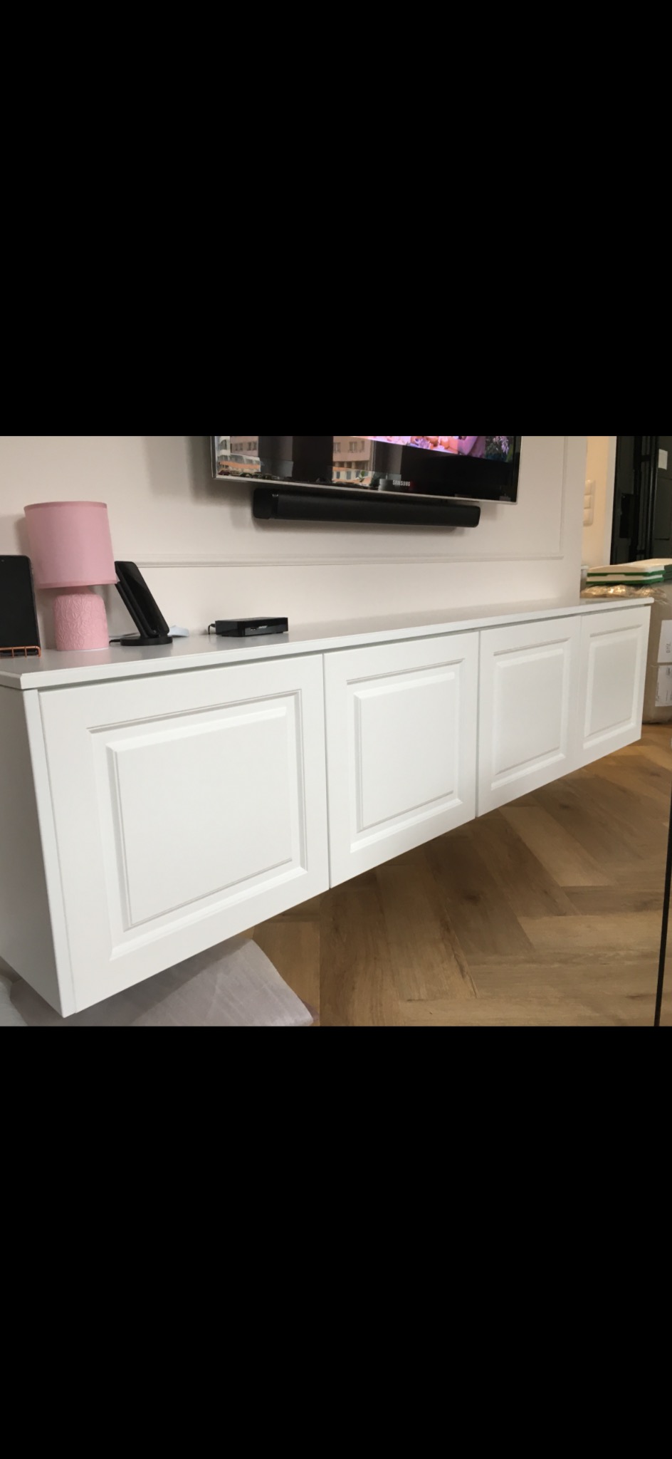 Biała, podwieszana szafka RTV z frezowanymi frontami, zamontowana pod telewizorem na ścianie. Parkiet ułożony w jodełkę. Element wystroju wnętrza.