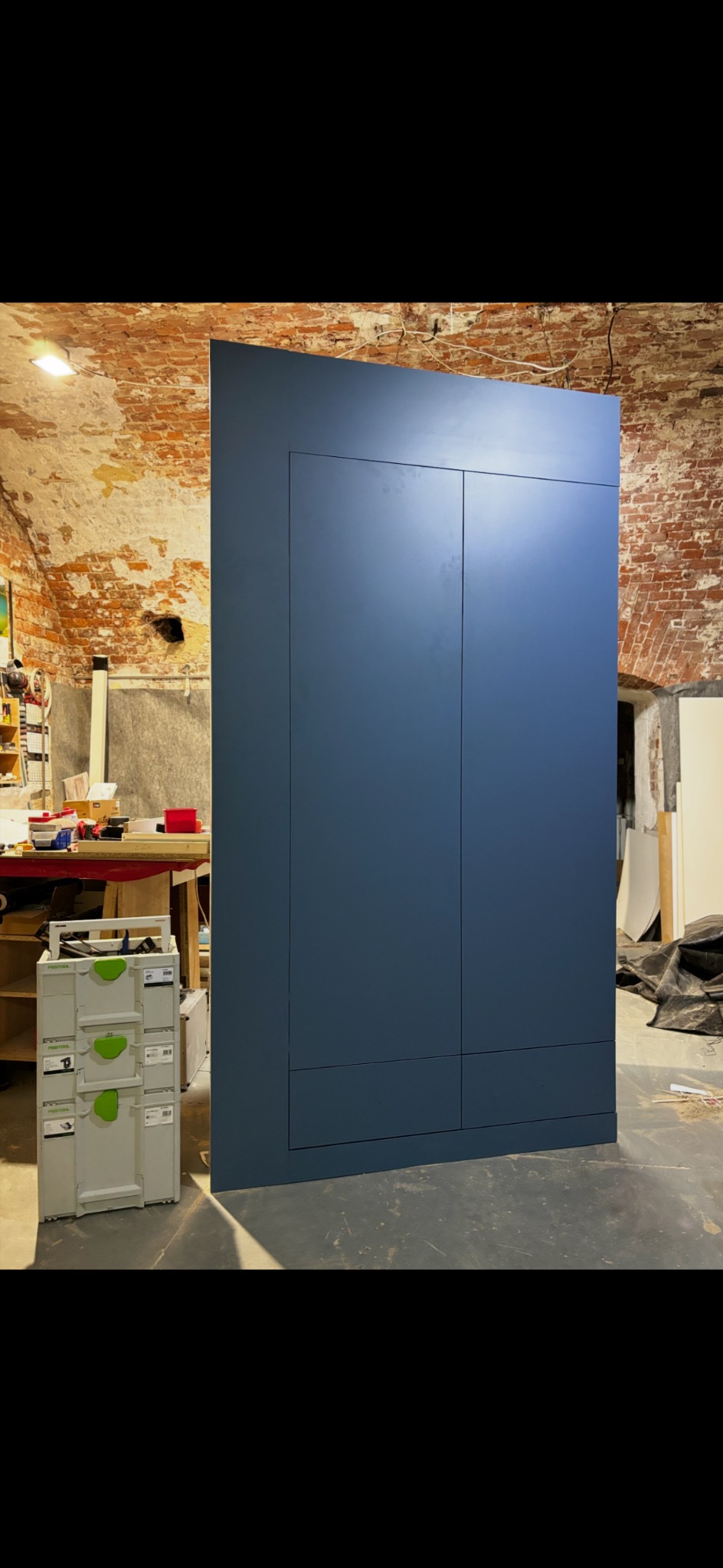 Granatowa szafa wnękowa z frezowanymi drzwiami, widoczna w surowym wnętrzu z ceglaną ścianą i skrzynką narzędziową Festool. Minimalistyczny design.