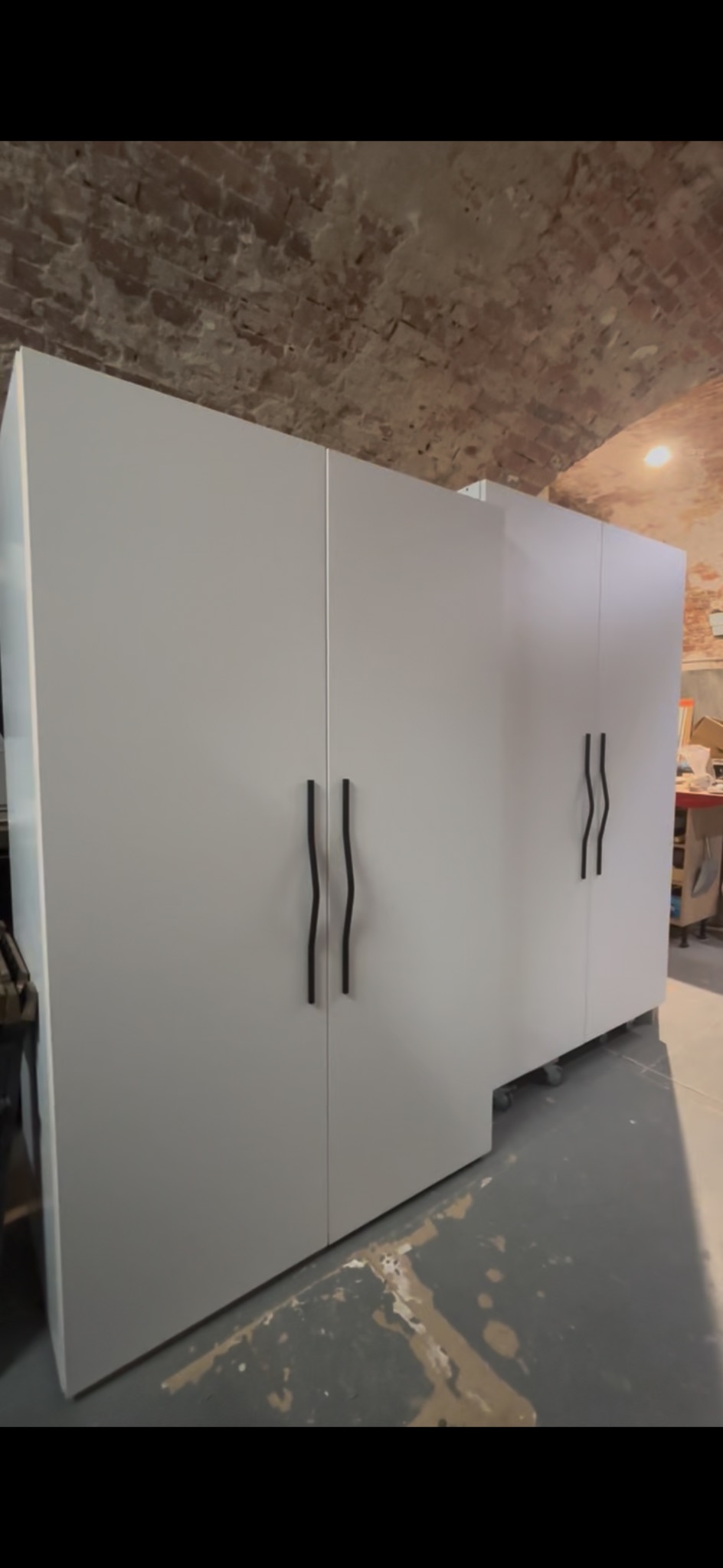 Biała szafa z czarnymi, falistymi uchwytami na tle ceglanego sufitu. Widoczne kółka u podstawy szafy. Nietypowe połączenie nowoczesności i surowego wnętrza.