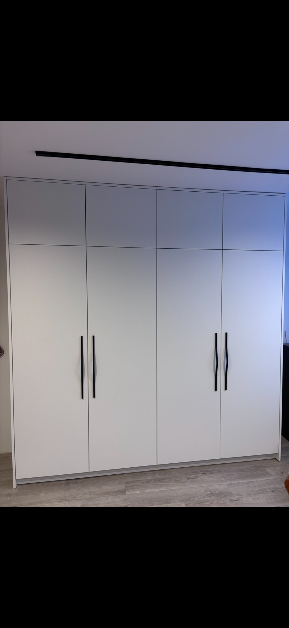 Biała, minimalistyczna szafa wnękowa z czterema drzwiami i długimi, czarnymi uchwytami, umieszczona na tle jasnej podłogi i sufitu z czarną listwą.