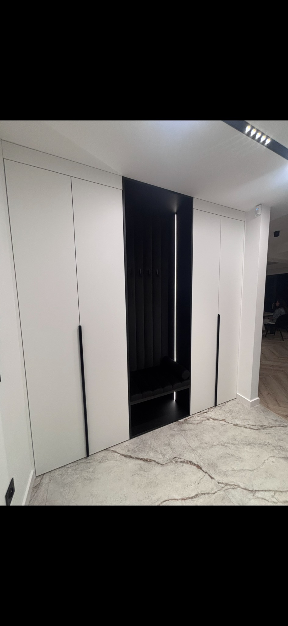 Biała szafa wnękowa z czarnym siedziskiem i oświetleniem LED, minimalistyczny design, marmurowa podłoga, nowoczesne wnętrze.