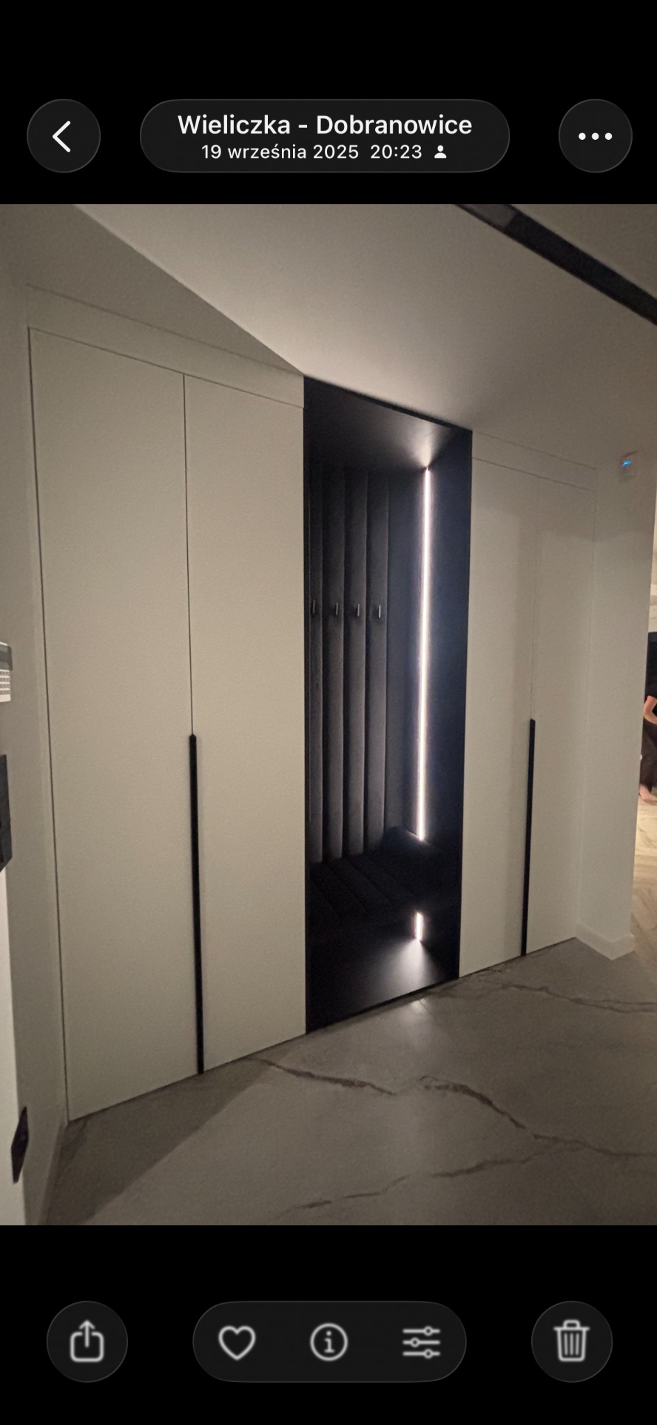 Nowoczesna szafa wnękowa z białymi frontami i czarnym, podświetlanym wnętrzem. Minimalistyczny design, długie, czarne uchwyty. Podłoga z szarego marmuru.