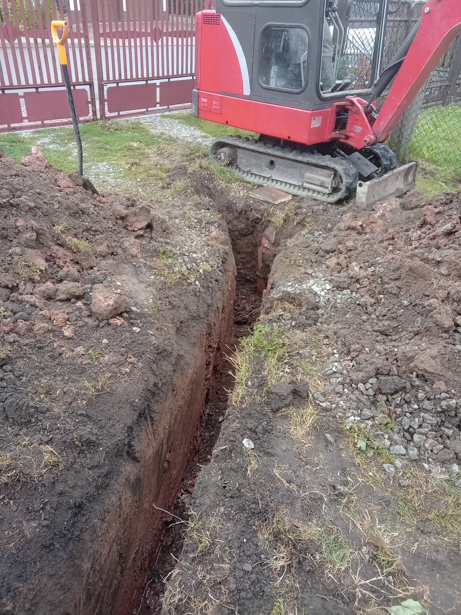 Wąski wykop w ziemi, obok czerwona mini koparka gąsienicowa i łopata z żółtą rączką przy metalowej bramie.