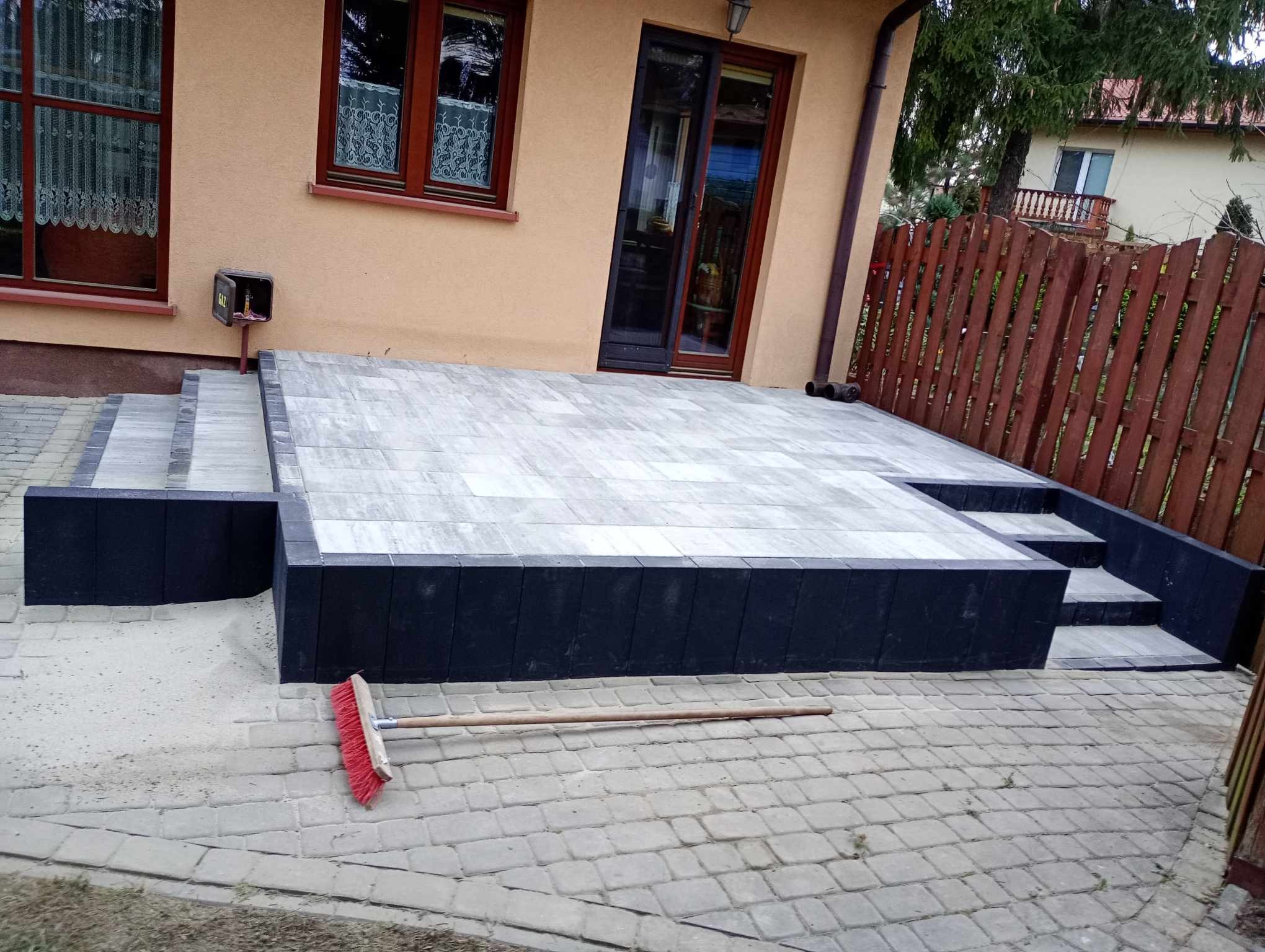 Wykończony taras z jasnoszarej kostki brukowej z ciemnymi obrzeżami, widoczne schody z kostki, czerwona szczotka leżąca na bruku, tło stanowi elewacja budynku i drewniany płot.