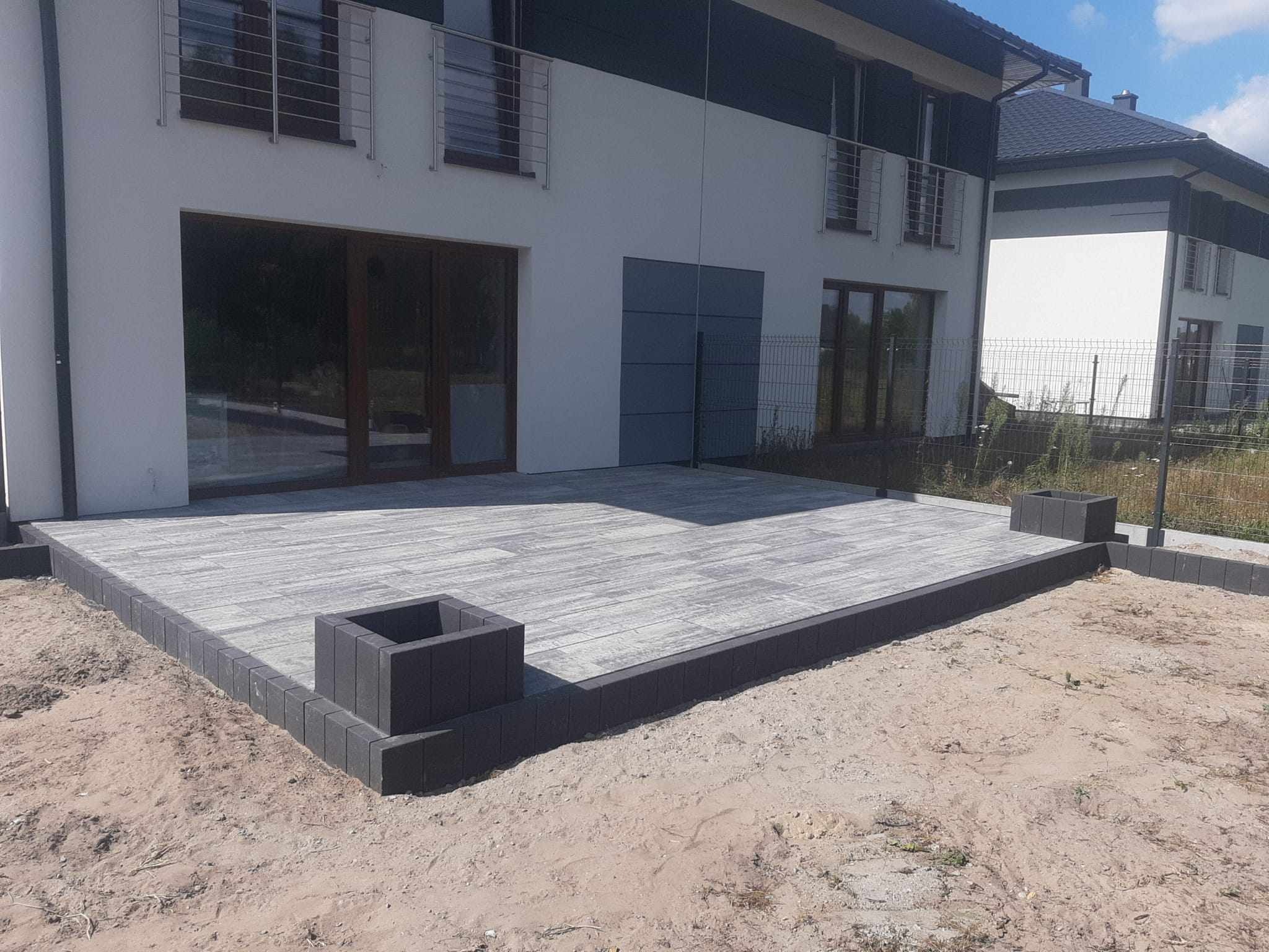 Nowoczesny taras z szarej kostki brukowej ułożony przy białym domu z ciemnymi elementami wykończeniowymi i szarą bramą garażową, widoczny fragment trawnika i ogrodzenia.