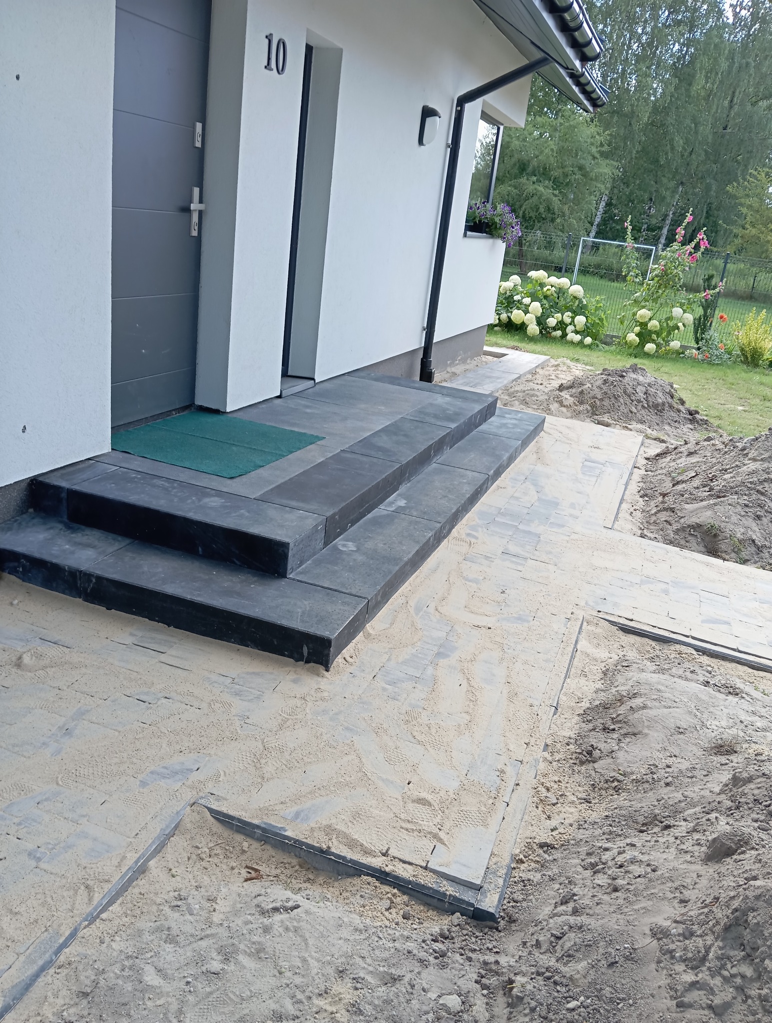 Nowe schody z ciemnoszarej kostki prowadzące do drzwi wejściowych, z fragmentem ułożonej ścieżki z kostki brukowej, zasypanej piaskiem, w tle trawnik i kwiaty.