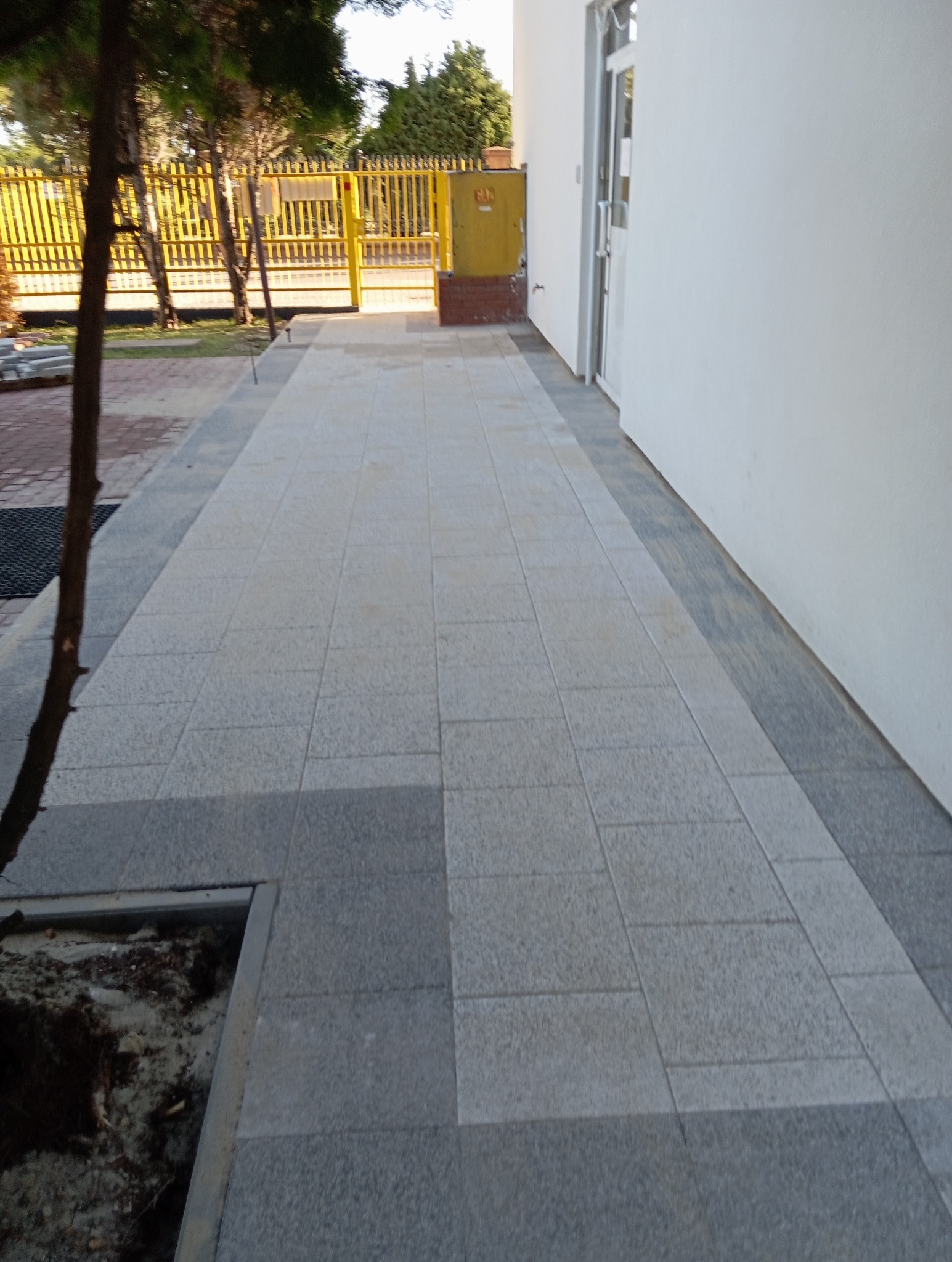 Ułożona jasnoszara kostka brukowa prowadząca do budynku z białymi ścianami i żółtą bramą w tle, widoczny fragment drzewa i donicy z ziemią po lewej stronie.