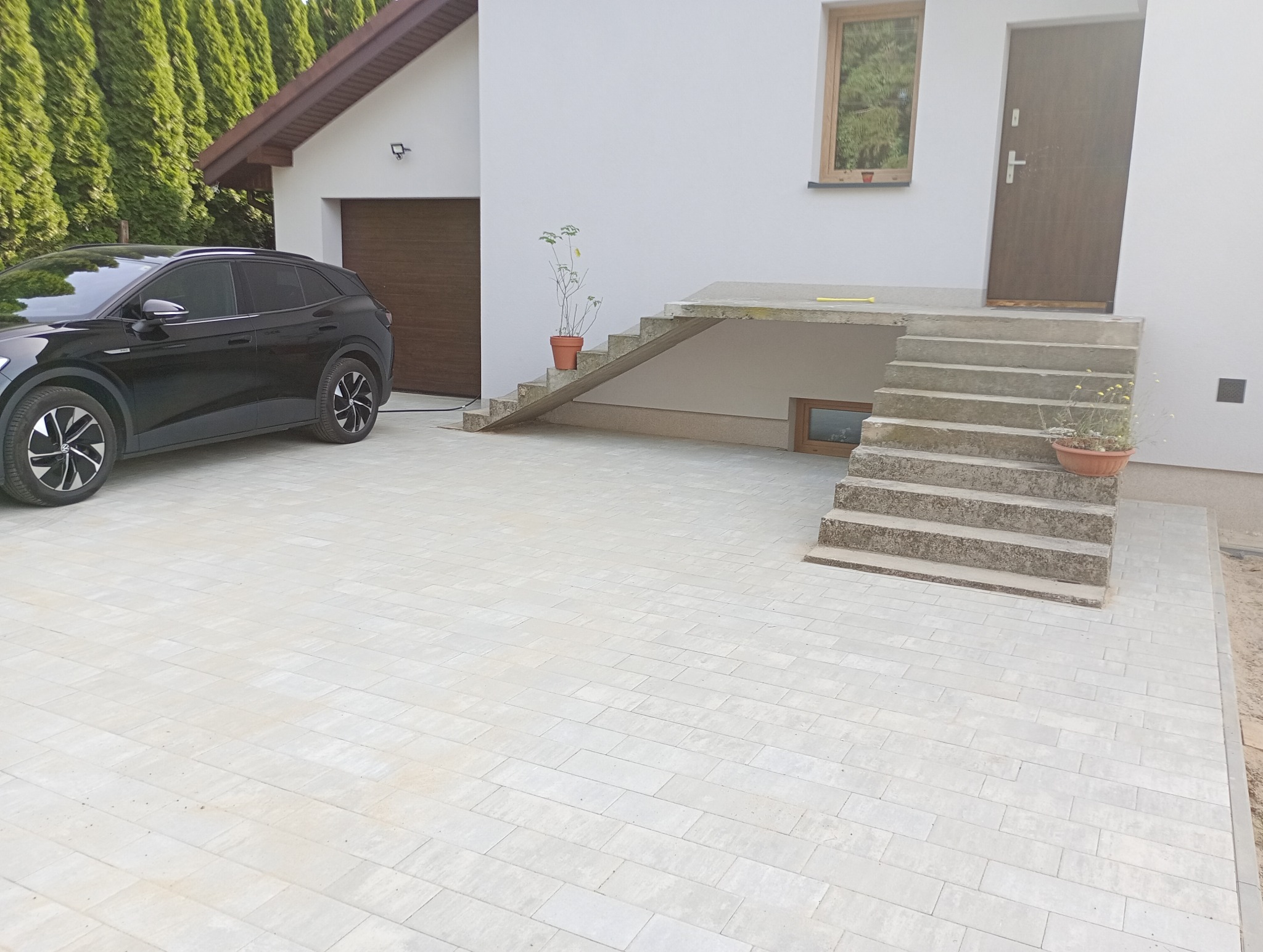 Jasnoszara kostka brukowa przed nowoczesnym domem z czarnym samochodem elektrycznym zaparkowanym obok, schody wejściowe z doniczkami po bokach.
