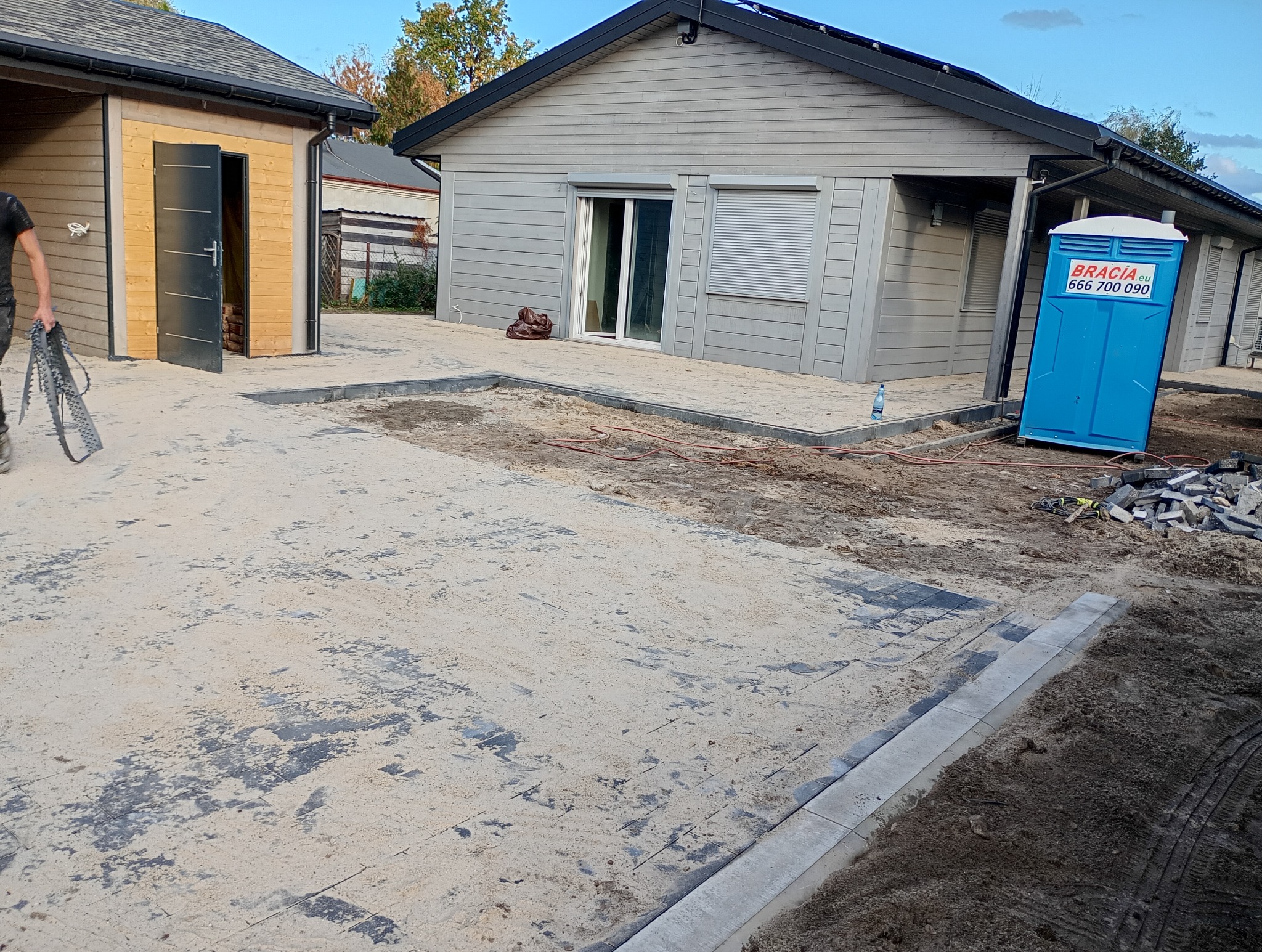 Układanie szarej kostki brukowej na posesji, widoczny częściowo ułożony teren, piasek, obrzeża, budynki w tle i przenośna toaleta.
