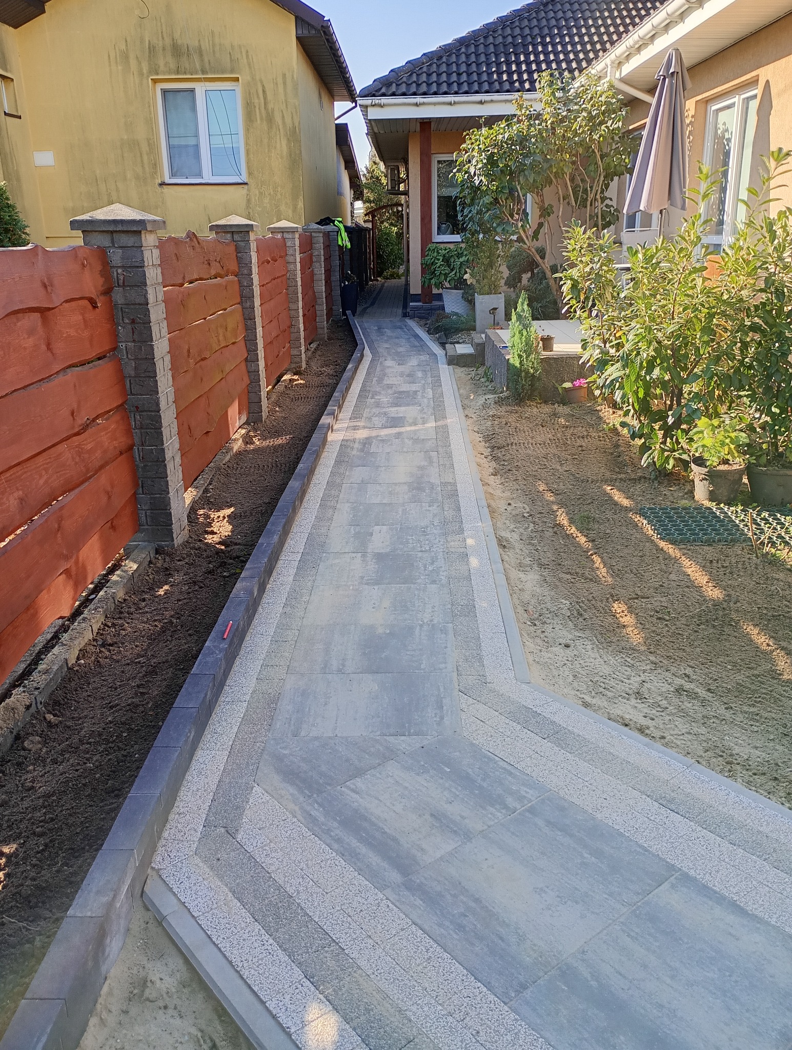 Długa, nowa ścieżka z szarych płyt betonowych z jasnymi obrzeżami, prowadząca wzdłuż drewnianego płotu z ceglanymi słupkami i rabaty z ziemią, pomiędzy dwoma budynkami.