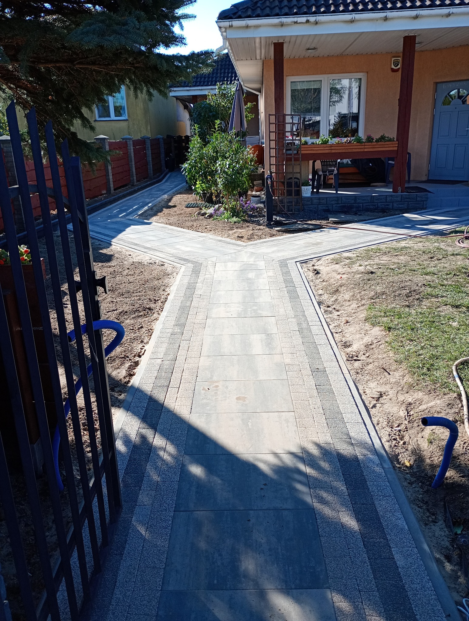 Ścieżka z szarych płyt betonowych z obramowaniem z kostki brukowej prowadząca do domu z drewnianym gankiem, widok zza metalowej furtki z niebieskim wężem ogrodowym w tle.