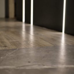 Wykończenia wnętrz od A do Z Tomasz Rzepka - Połączenie podłogi z paneli drewnopodobnych z szarą płytką imitującą kamień, oświetlona pionowymi pasami światła w ciemnym pomieszczeniu.