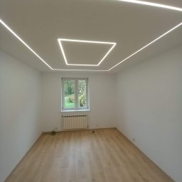 Wykończenia wnętrz od A do Z Tomasz Rzepka - Pusty pokój z panelami podłogowymi w odcieniach jasnego drewna i nowoczesnym oświetleniem sufitowym w postaci białych, kwadratowych LED-owych linii.