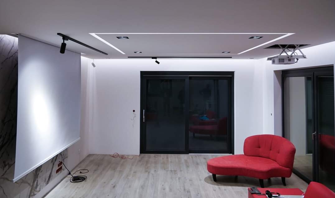 Nowoczesny salon z projektorem zamontowanym na suficie, ekranem zwijanym na ścianie z imitacją marmuru, oświetleniem LED wpuszczonym w sufit i czerwonym szezlongiem.