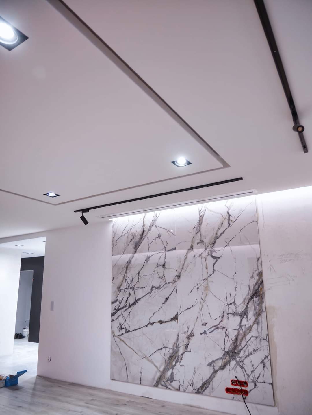 Nowoczesne wnętrze z podświetlaną ścianą z płytek imitujących marmur, oświetleniem szynowym i podwieszanym sufitem z wbudowanymi punktami świetlnymi.