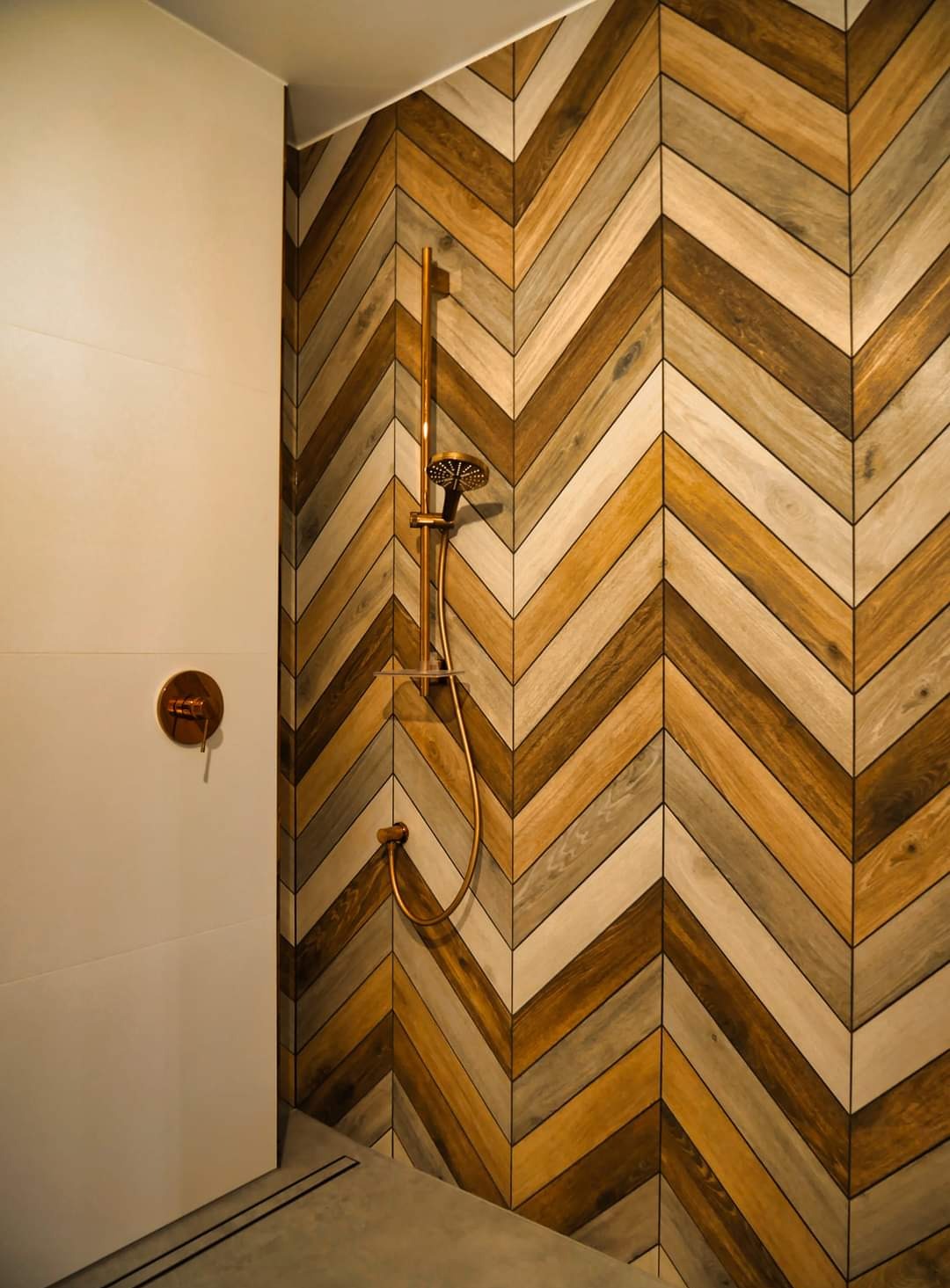 Nowoczesna kabina prysznicowa z drewnopodobnymi płytkami ułożonymi w jodełkę i miedzianą armaturą.