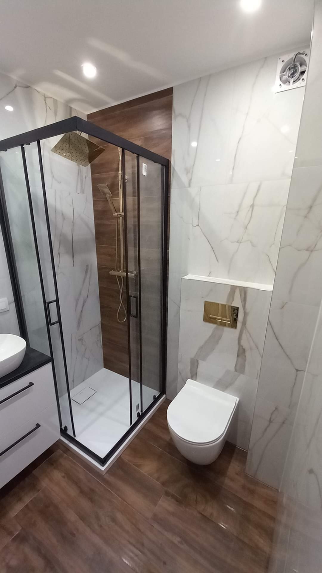Nowoczesna łazienka z kabiną prysznicową typu walk-in z czarnymi profilami, armaturą w kolorze złotym, marmurowymi płytkami i drewnopodobną podłogą ułożoną w jodełkę.