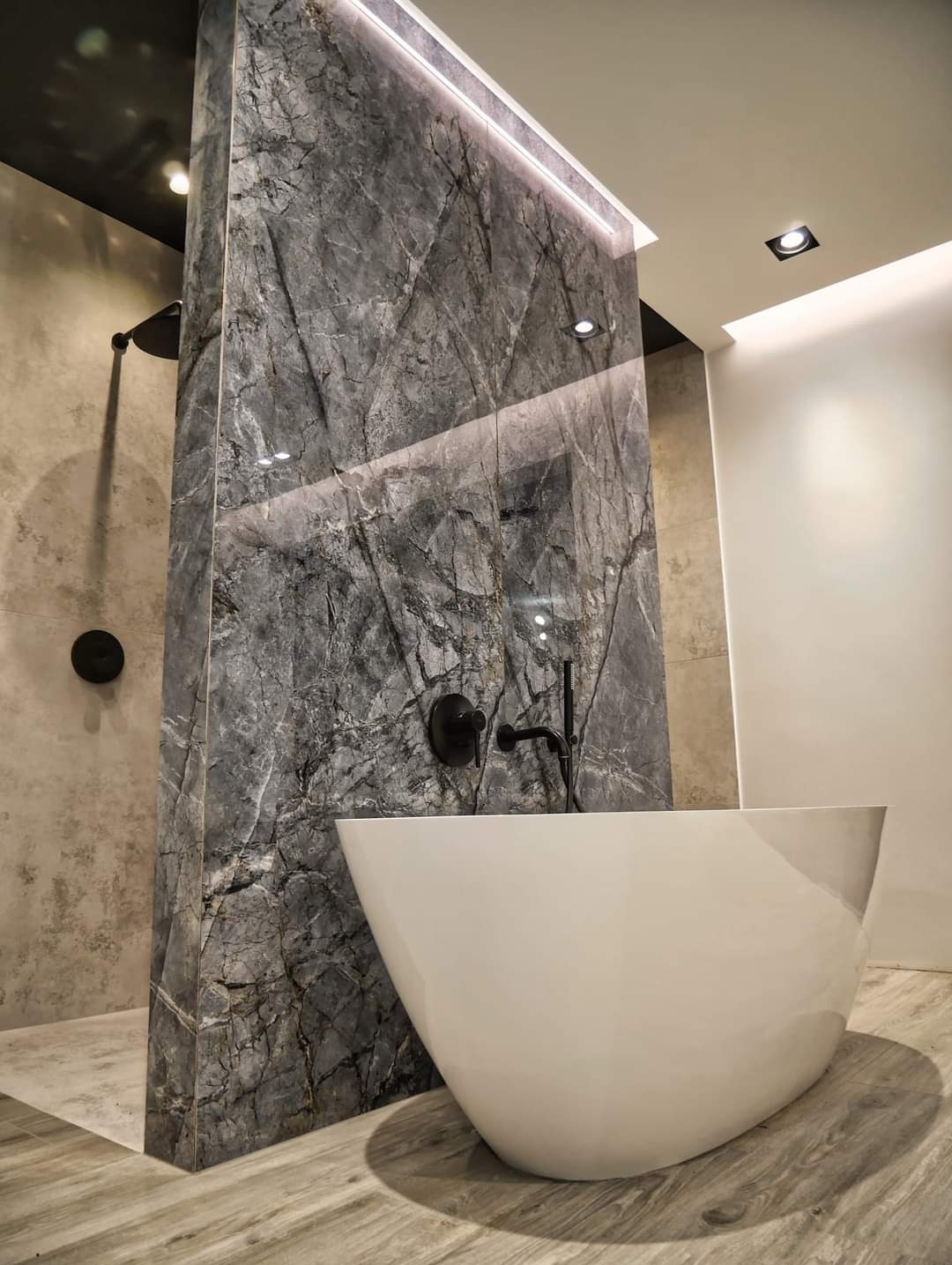 Nowoczesna łazienka z wolnostojącą białą wanną, ścianą z szarego marmuru z podświetleniem LED i czarną armaturą.