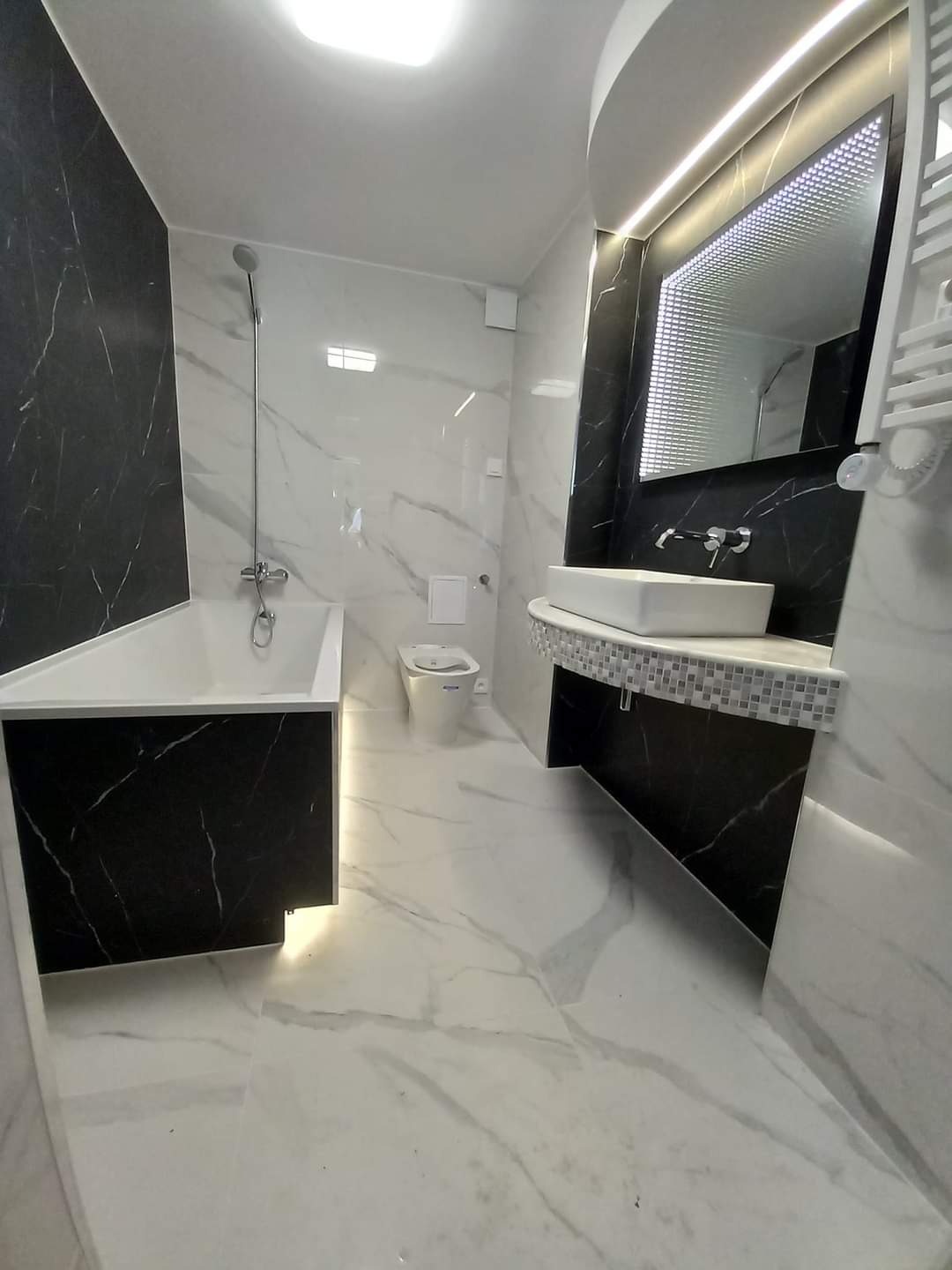 Nowoczesna łazienka z czarno-białymi marmurkowymi płytkami, podświetlanym lustrem i minimalistyczną armaturą, widok na wannę, toaletę i umywalkę.