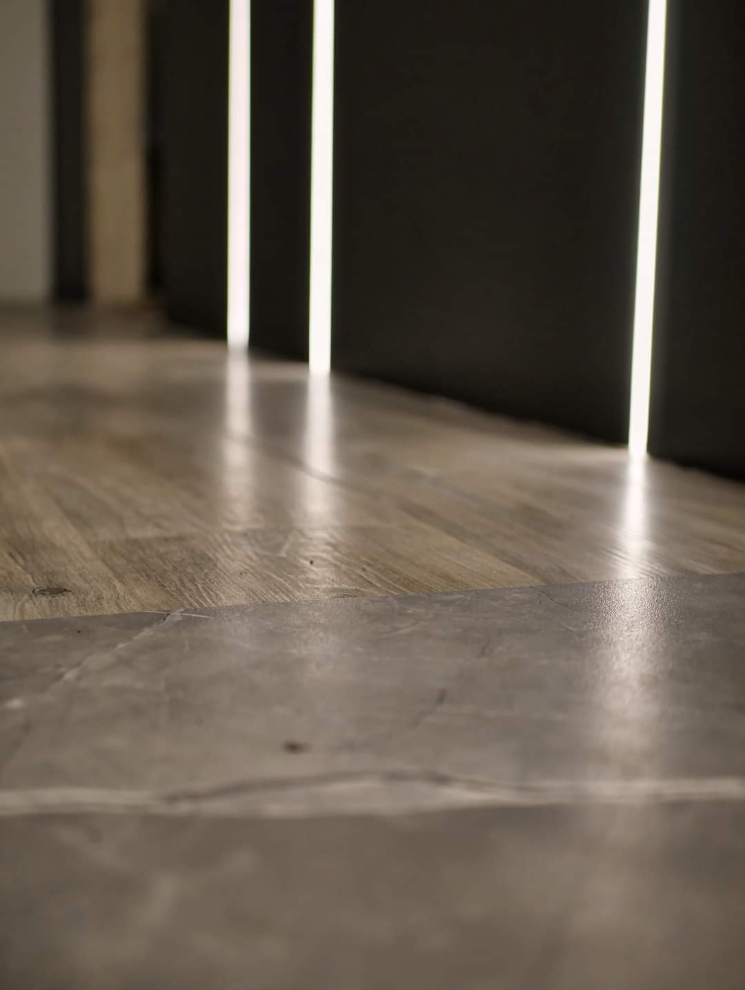 Połączenie podłogi z paneli drewnopodobnych z szarą płytką imitującą kamień, oświetlona pionowymi pasami światła w ciemnym pomieszczeniu.