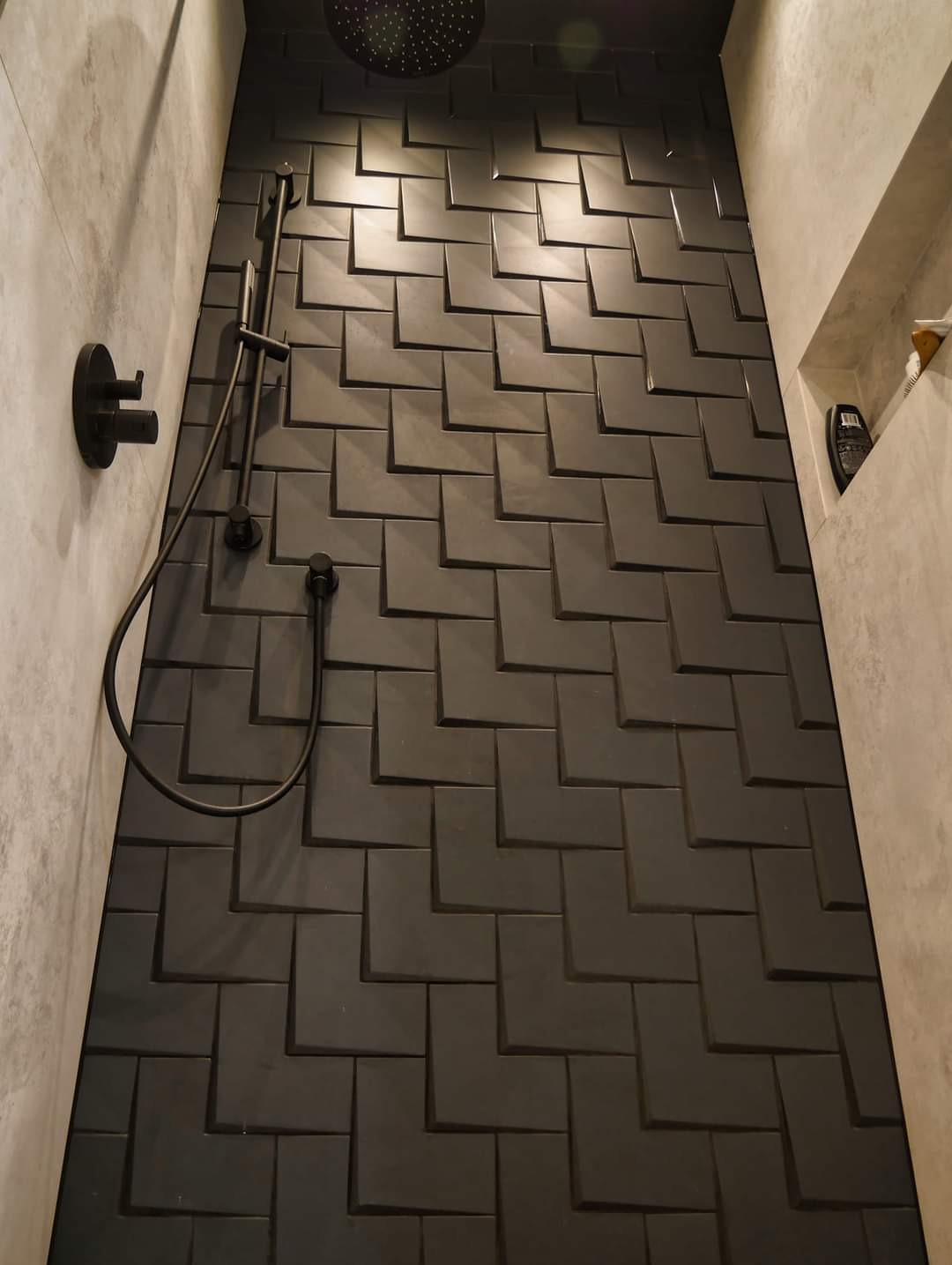 Nowoczesna kabina prysznicowa z czarnymi, trójwymiarowymi płytkami ułożonymi w jodełkę, deszczownicą i minimalistyczną armaturą w kolorze czarnym, oraz półką wnękową z kosmetykami.