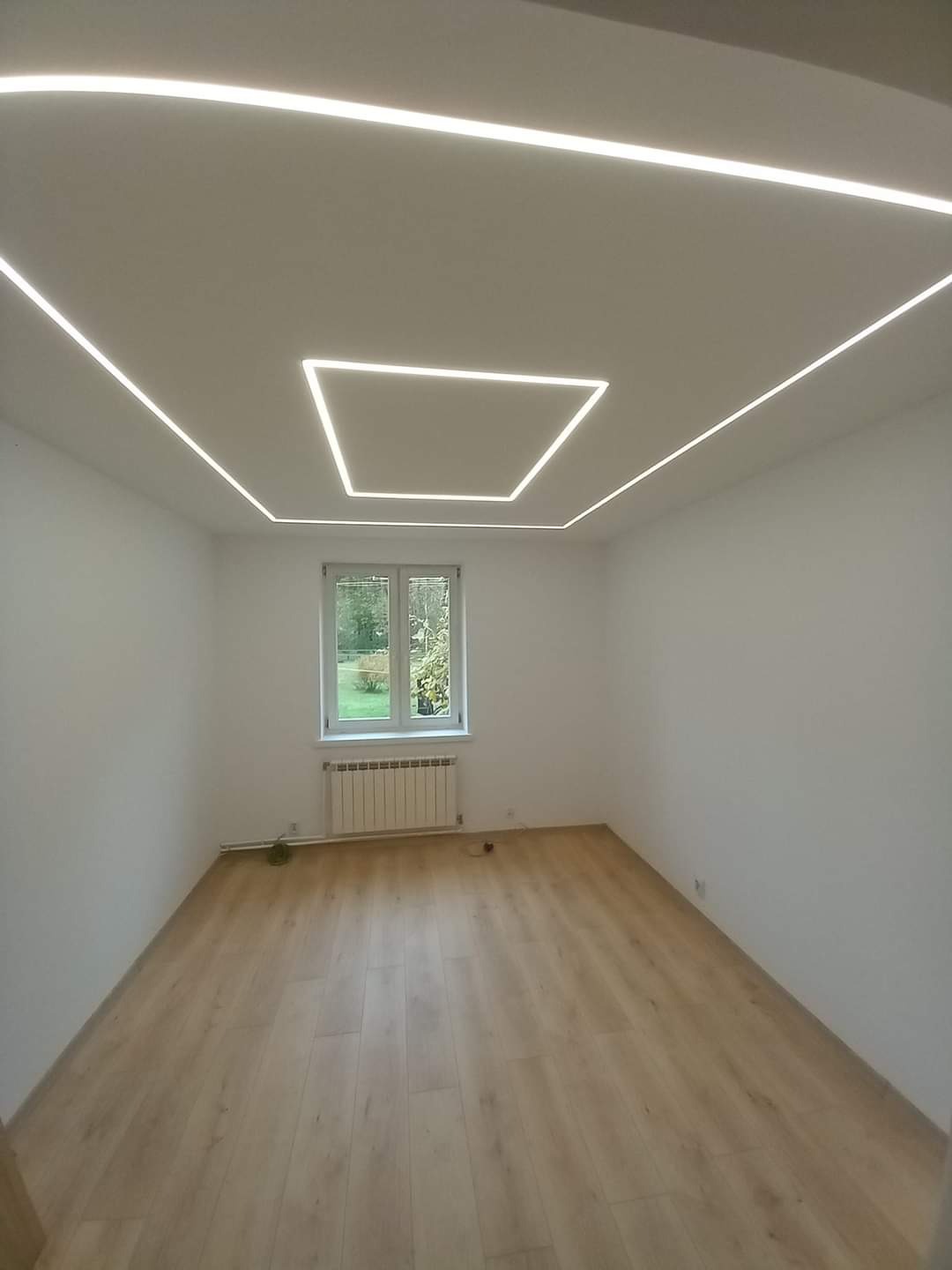 Pusty pokój z panelami podłogowymi w odcieniach jasnego drewna i nowoczesnym oświetleniem sufitowym w postaci białych, kwadratowych LED-owych linii.