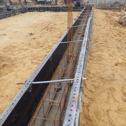 GRACZTEAM - Zbrojenie ławy fundamentowej w szalunkach systemowych na piaszczystym podłożu, widoczne pionowe pręty i poziome zbrojenie, w tle dom z panelami słonecznymi.