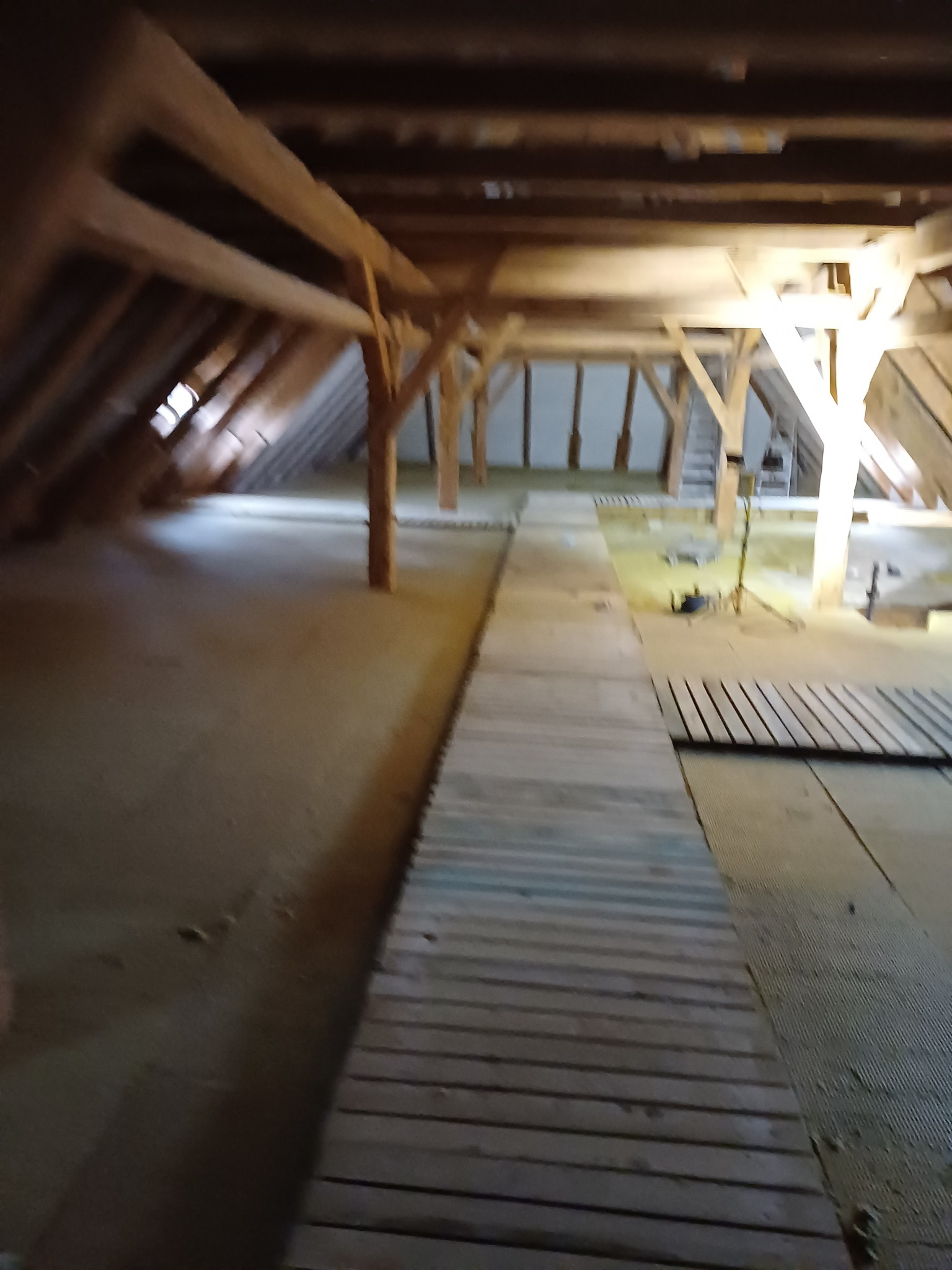 Poddasze z drewnianą konstrukcją więźby dachowej, częściowo ocieplone, z prowizorycznym chodnikiem z desek, widoczny sprzęt budowlany i drabina.