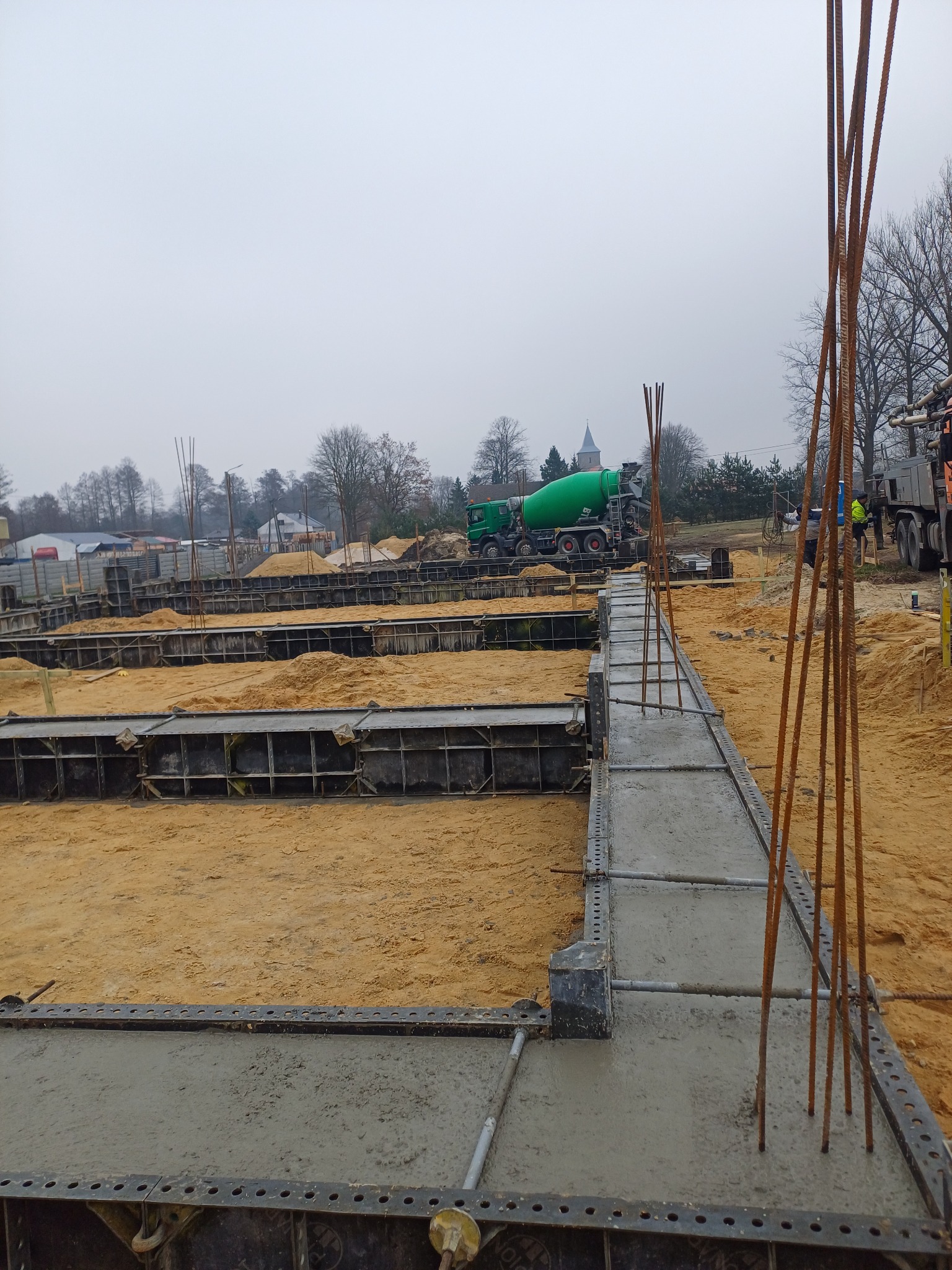 Wylany beton w szalunkach z wystającymi prętami zbrojeniowymi, w tle zielona betoniarka i kościół w oddali.