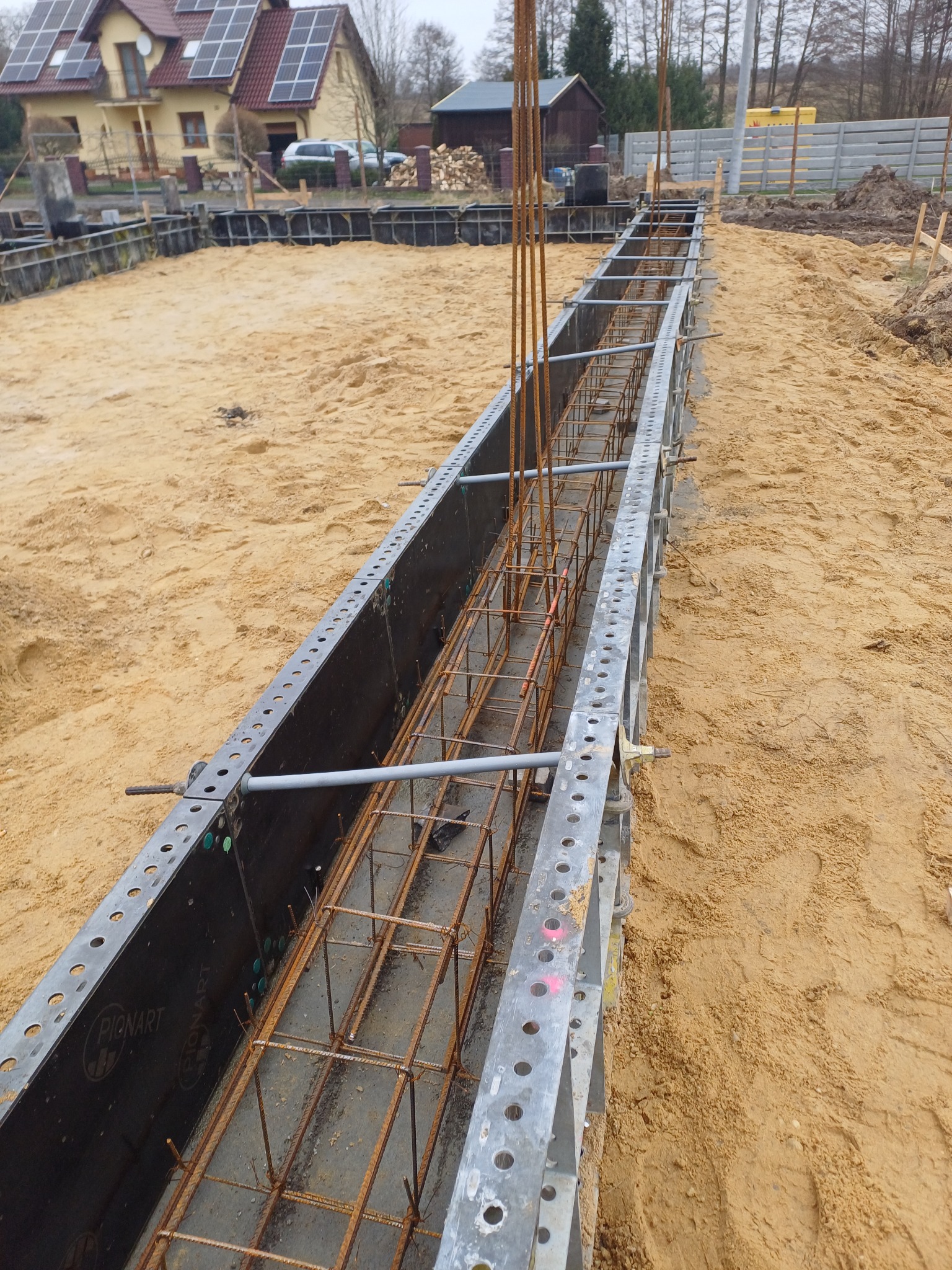 Zbrojenie ławy fundamentowej w szalunkach systemowych na piaszczystym podłożu, widoczne pionowe pręty i poziome zbrojenie, w tle dom z panelami słonecznymi.