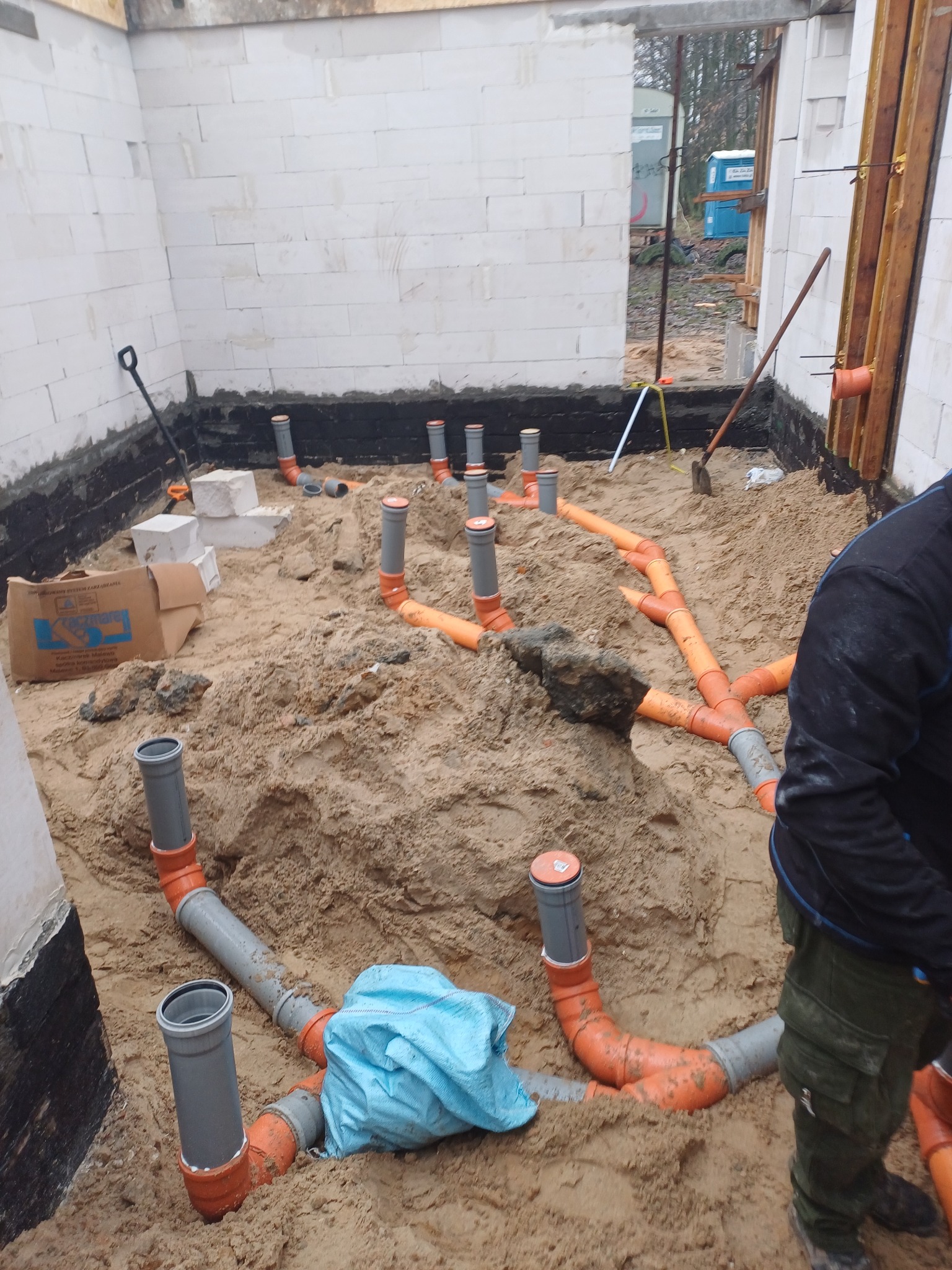 Instalacja kanalizacyjna w piasku, widoczne rury pomarańczowe i szare, ściany z białych bloczków, przygotowanie do wylewki w budynku w stanie surowym otwartym.