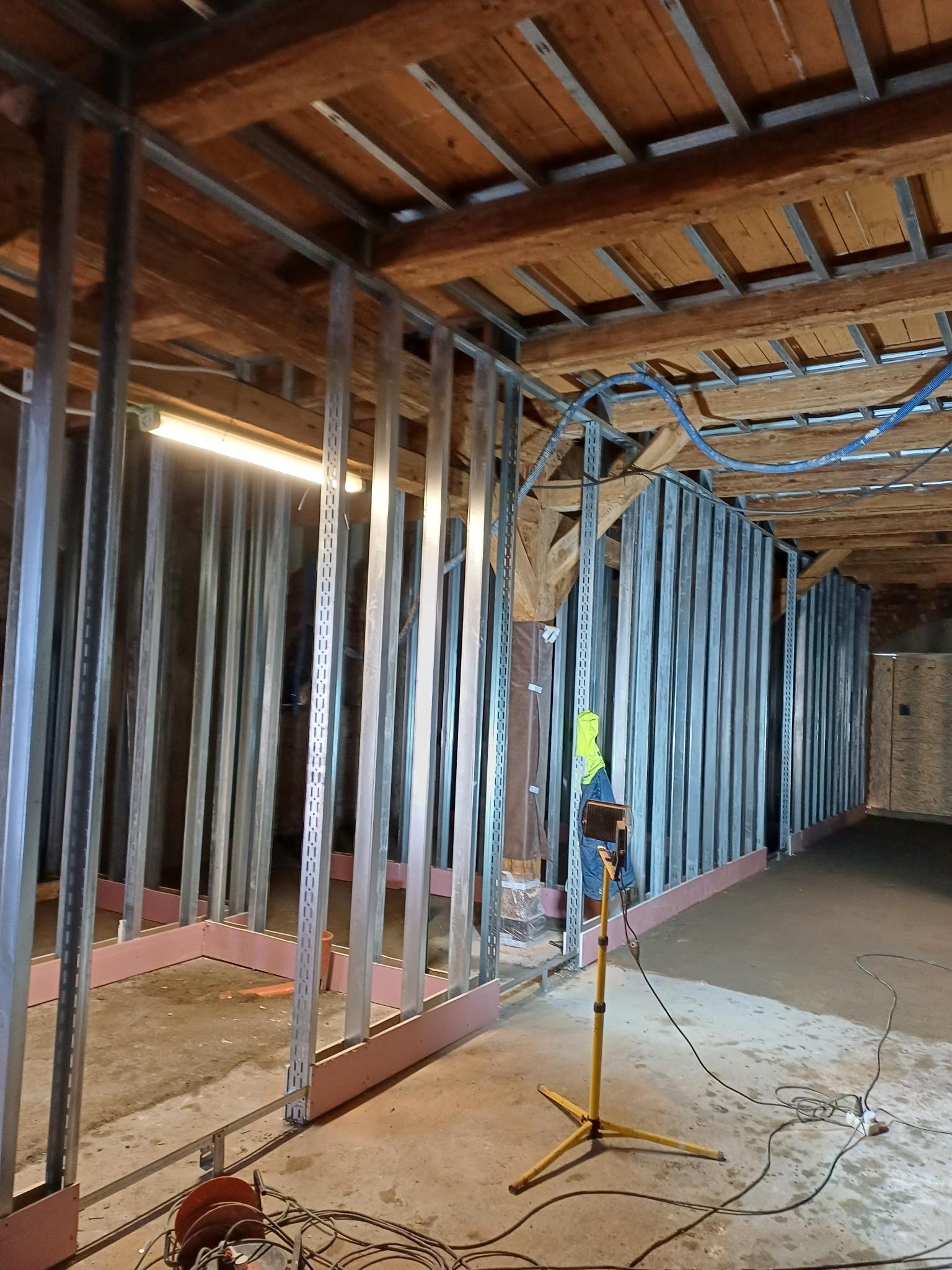 Wnętrze w trakcie budowy z metalową konstrukcją ścian działowych, widoczne drewniane belki stropowe, różowe elementy izolacyjne przy podłodze, oświetlenie budowlane na statywie i plątanina kabli.