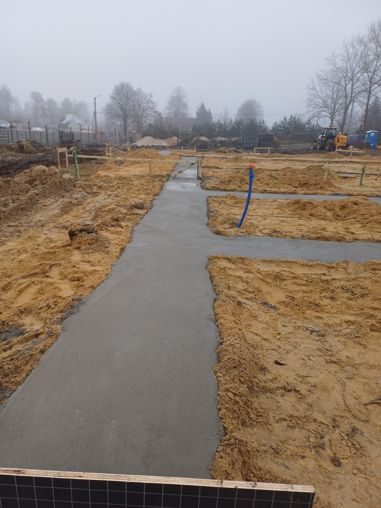 Świeżo wylany fundament z betonu na placu budowy, widoczne drewniane szalunki i wystająca niebieska rura instalacyjna w pochmurny dzień.
