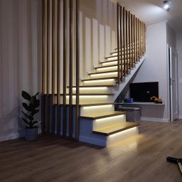 Usługi Stolarskie Paweł Jabłoński - Nowoczesne schody z drewnianymi stopniami podświetlonymi ledami, z drewnianą ażurową balustradą, obok doniczka z rośliną, w tle pokój dziecięcy z tablicą i zabawkami.