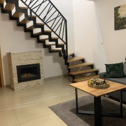 Usługi Stolarskie Paweł Jabłoński - Nowoczesne schody z drewnianymi stopniami i czarną metalową balustradą w jasnym wnętrzu z kominkiem i drewnianym stolikiem kawowym.