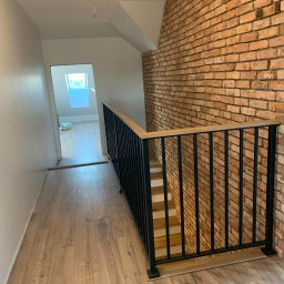 Usługi Stolarskie Paweł Jabłoński - Nowoczesne schody z drewnianymi stopniami i czarną metalową balustradą, widok z góry na drewnianą podłogę, ściana z cegły w tle.