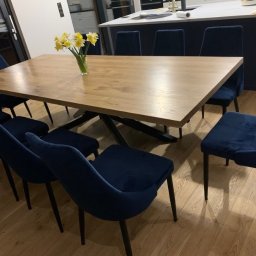 Usługi Stolarskie Paweł Jabłoński - Stół z blatem z jasnego drewna o geometrycznej podstawie z czarnych metalowych elementów, otoczony granatowymi krzesłami z weluru, na podłodze z drewnianych paneli, z bukietem żółtych narcyzów...