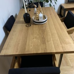 Usługi Stolarskie Paweł Jabłoński - Jasny, dębowy blat stołu z dekoracjami świątecznymi, czarnymi świecznikami i krzesłami z ciemną tapicerką, ustawiony w jasnym wnętrzu z parkietem i roślinami.