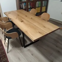 Stół z jasnego drewna z widocznymi sękami, na czarnych, metalowych nogach, otoczony czterema krzesłami z drewnianymi oparciami i czarnymi siedziskami, ustawiony na jasnej podłodze z panelami,...