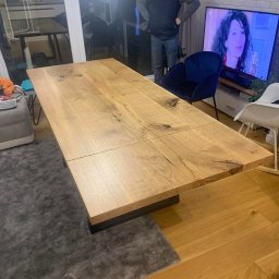 Usługi Stolarskie Paweł Jabłoński - Rozkładany stół z blatem z jasnego drewna o widocznej strukturze słojów, osadzony na czarnej, minimalistycznej podstawie, w tle mężczyzna i wnętrze salonu z telewizorem.