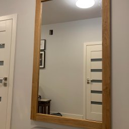 Usługi Stolarskie Paweł Jabłoński - Duże lustro w drewnianej ramie z widocznym odbiciem wnętrza, w tle białe drzwi i lampa sufitowa, pod lustrem szara tapicerowana ławka.