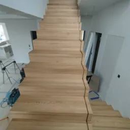 Wysokie, drewniane schody o jasnym odcieniu drewna, prowadzące do góry w nowoczesnym wnętrzu, widoczne narzędzia i elementy wykończeniowe sugerują trwający montaż.