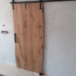 Usługi Stolarskie Paweł Jabłoński - Surowe drewniane drzwi przesuwne z czarnym okuciem na tle surowej ściany, widoczne instalacje elektryczne.