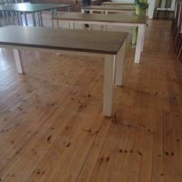 Usługi Stolarskie Paweł Jabłoński - Sala z drewnianą podłogą, stołami z jasnymi nogami i blatami w kolorze drewna oraz rzędem krzeseł ustawionych wzdłuż dłuższego stołu przykrytego białym obrusem.