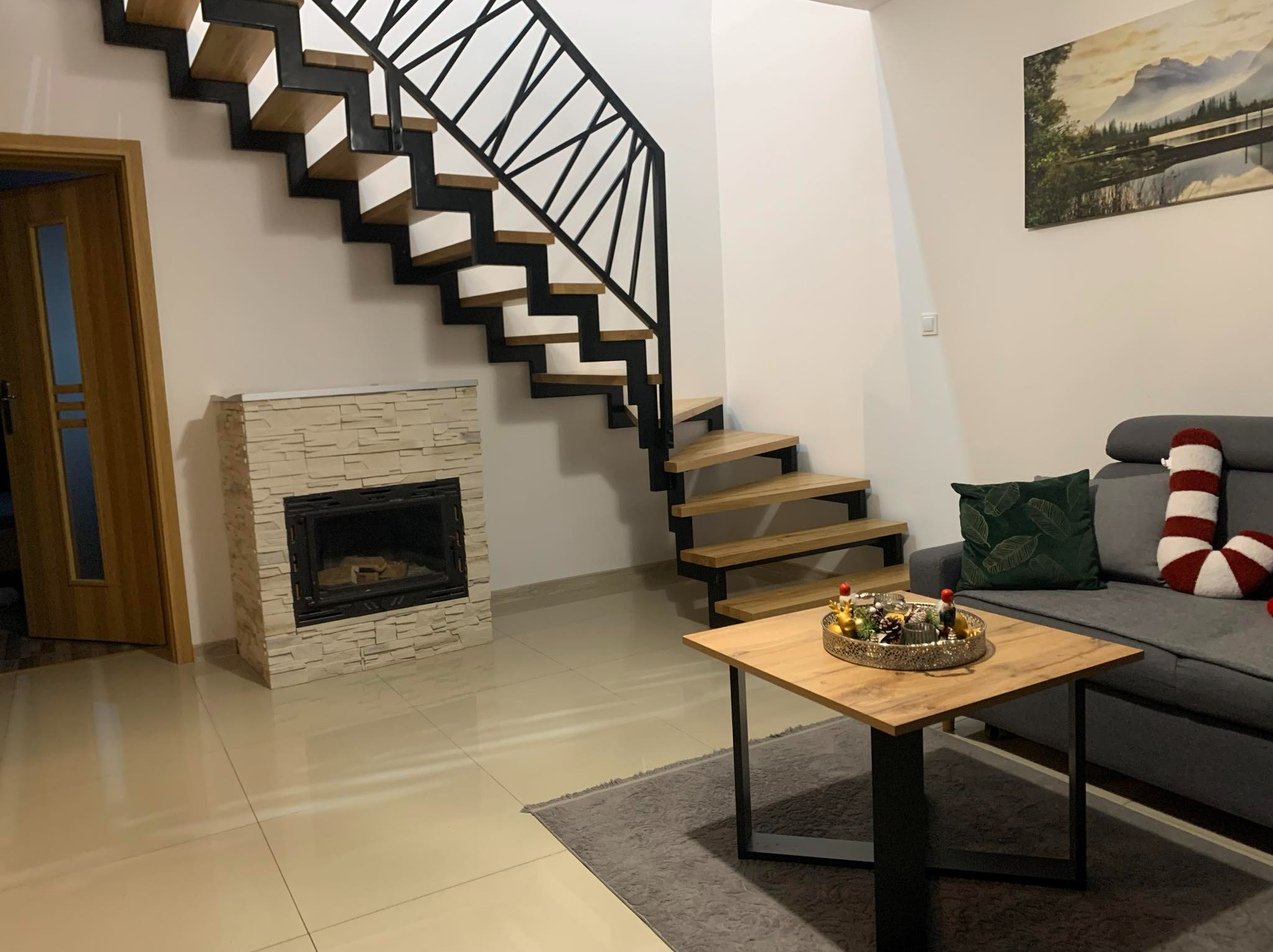 Nowoczesne schody z drewnianymi stopniami i czarną metalową balustradą w jasnym wnętrzu z kominkiem i drewnianym stolikiem kawowym.