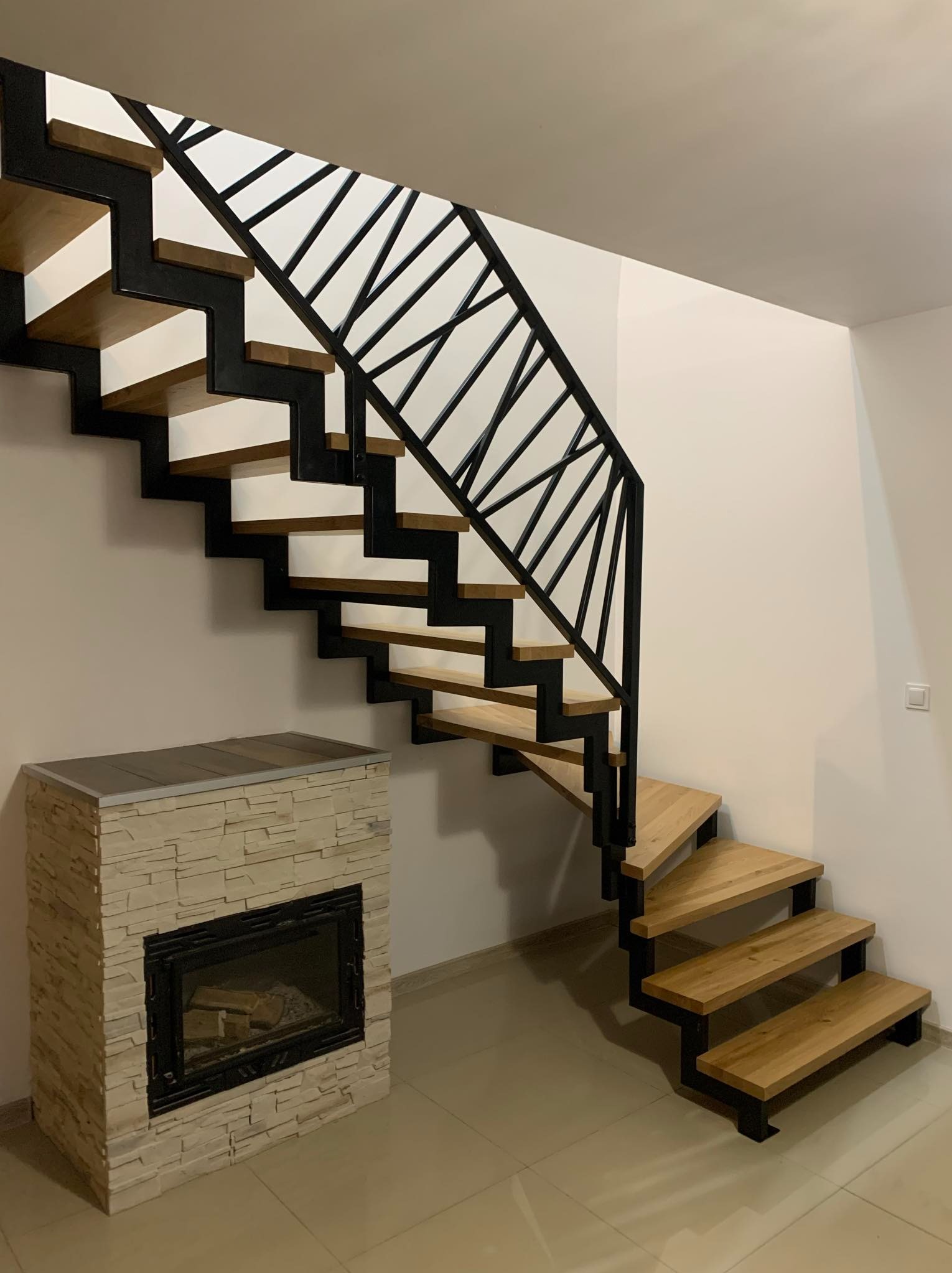 Nowoczesne schody z drewnianymi stopniami i czarną, geometryczną balustradą, umieszczone w jasnym wnętrzu obok obudowanego kominka.
