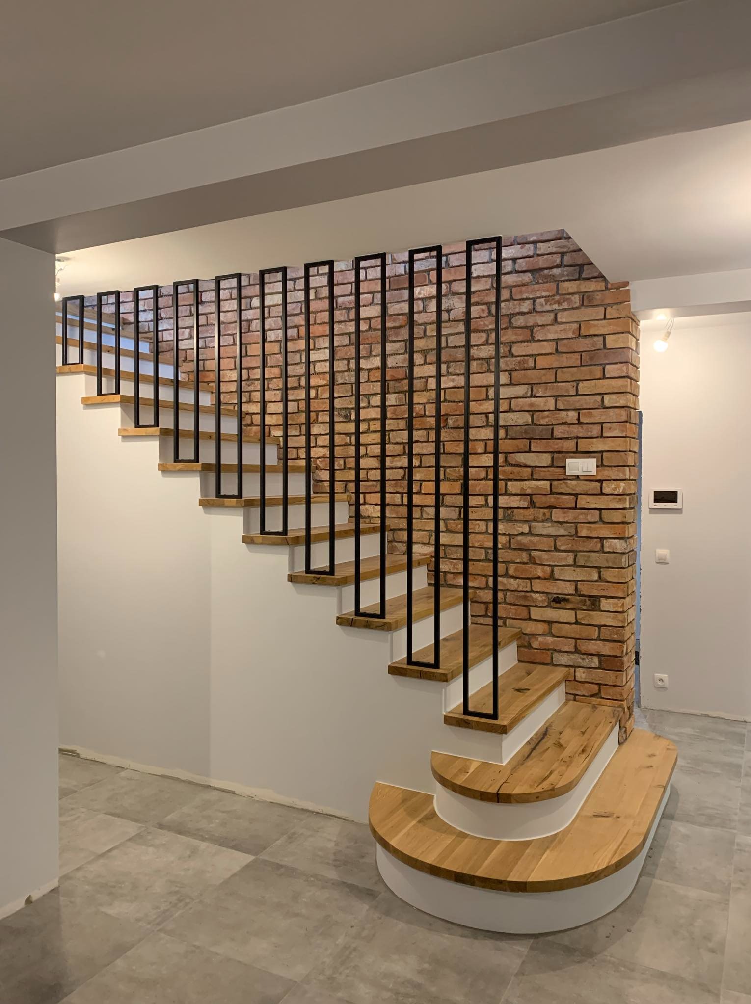 Nowoczesne schody drewniane z czarną, minimalistyczną balustradą na tle ściany z czerwonej cegły, oświetlone punktowymi lampami.