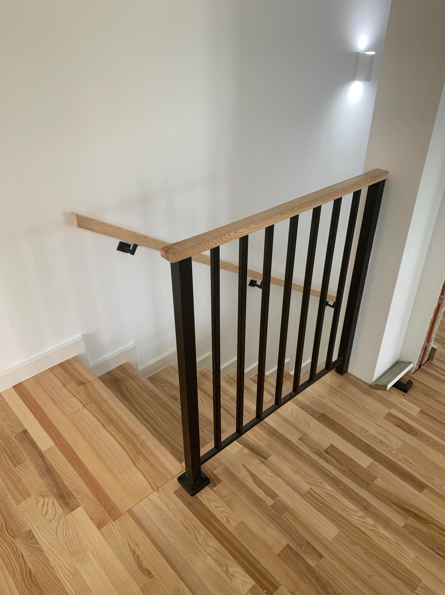 Nowoczesne schody z drewnianymi stopniami i czarną metalową balustradą z drewnianym pochwytem, oświetlone kinkietem ściennym.