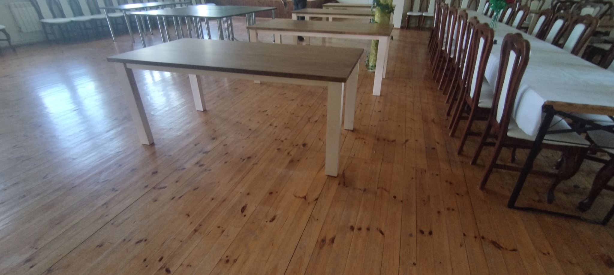 Sala z drewnianą podłogą, stołami z jasnymi nogami i blatami w kolorze drewna oraz rzędem krzeseł ustawionych wzdłuż dłuższego stołu przykrytego białym obrusem.